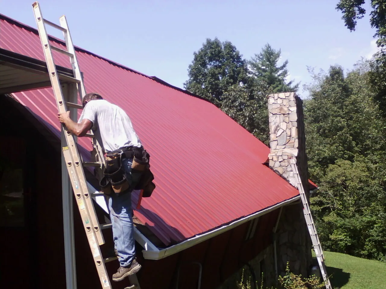 Roofer on a ladder beside a red metal roof and stone chimney — Soffit & Fascia Repair by Roofing Brothers in Priest River